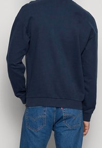 Persona che indossa una felpa blu navy e jeans in denim blu, vista da dietro su uno sfondo chiaro uniforme.
