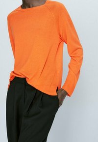 Oranger Pullover mit langen Ärmeln, rundem Ausschnitt, Raglanärmeln und lockerer Passform, kombiniert mit schwarzen, gefältelten Hosen.
