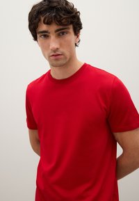 Giovane con capelli ricci e scuri che indossa una maglietta rossa a maniche corte e girocollo, su uno sfondo neutro chiaro.