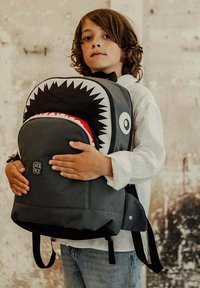 Gray backpack with shark face design, black teeth, and red accents. Features adjustable straps and a logo patch on the front.
