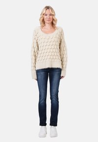 Cream-colored sweater with a textured, scale-like pattern, paired with dark blue jeans and white sneakers. Casual fit, long sleeves, crew neck.