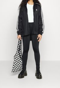 Svart zip-up jacka med vita ränder, svarta leggings, svarta kängor och en svart och vit rutig axelväska.