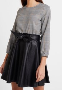 Haut à manches longues à motif à carreaux gris avec des accents marron, associé à une jupe en similicuir noir avec un détail noué à la taille.