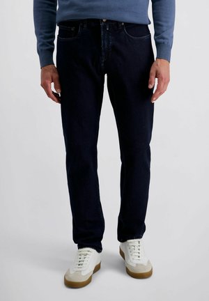 Mann trägt dunkelblaue Jeans, weiße Sneakers mit Gummisohlen und ein blaues Langarmshirt und steht vor einem schlichten Hintergrund.