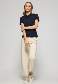 T-shirt polo bleu marine à manches courtes, col plat et logo rouge ; associé à un pantalon beige clair et des baskets beiges, affichant une coupe décontractée.