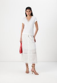 White v-neck t-shirt with lace trim, paired with a long white skirt featuring eyelet patterns and front buttons, complemented by a red bag.