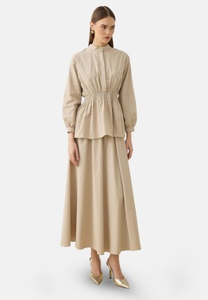 Kvinde iført en beige langærmet top med smukt tilpasset talje og matchende lang nederdel med guld højhælede sko, stående mod en ensfarvet baggrund.
