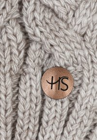 Knit sweater in light beige, featuring a textured cable pattern. Includes a round metallic logo with black lettering.
