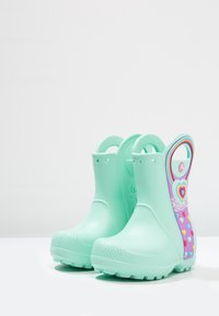 Bottes de pluie en caoutchouc vert menthe avec des semelles texturées, présentant un motif arc-en-ciel et cœurs d'un côté et une poignée pour un enfilage facile.