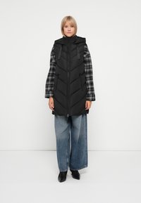 Kvinne i en svart polstret uten ermer hettevest over en svart og hvit rutete skjorte, vide blå jeans og svarte spisse sko.