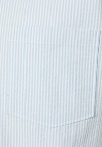 Un tessuto a righe blu chiaro presenta una tasca a toppa. La texture appare liscia con cuciture diritte lungo i bordi.