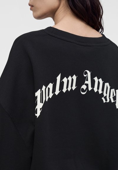 Γυναίκα που φοράει μαύρο φούτερ με "palm Angels" σε μεγάλα λευκά γοτθικά γράμματα στην πλάτη, κοιτάζοντας μακριά από την κάμερα.