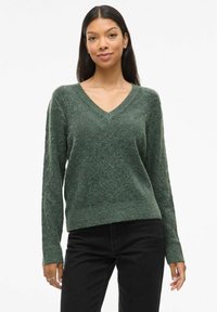Pull vert en V avec manches longues, texture douce et coupe décontractée. Associé à un jean noir, présenté sur un fond neutre.