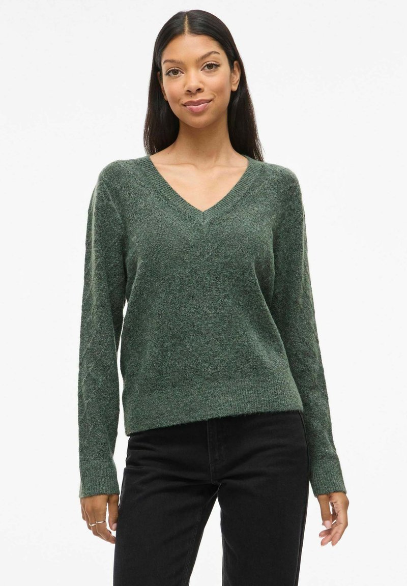 Pull vert en V avec manches longues, texture douce et coupe décontractée. Associé à un jean noir, présenté sur un fond neutre.