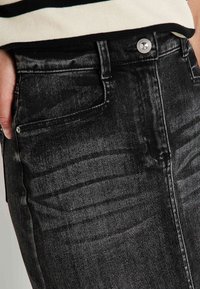 Primer plano de una persona que lleva jeans negros desgastados con botón plateado, con la mano resting en el bolsillo delantero y una camiseta blanca con ribete negro.