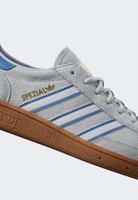 Pelēkie ādas sporta apavi ar ziliem akcentiem un trim baltām svītrām. Tajos ir teksturēta gumijas zole un zelta "SPEZIAL" zīmols.