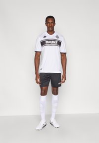 Hvid fodboldtrøje med sorte accenter og "MAXBET"-logo, kombineret med sorte shorts, hvide sokker og sorte fodboldstøvler. Enkel design og pasform.