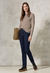 Maglione a maglia beige con maniche lunghe, jeans blu scuro e stivali neri alla caviglia con una suola spessa color cuoio. Tiene un giubbotto imbottito verde.