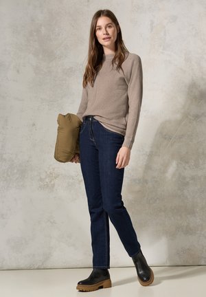 Pull en tricot beige à manches longues, jean bleu foncé et bottes noires à cheville avec une semelle épaisse couleur tan. Tient une veste rembourrée verte.