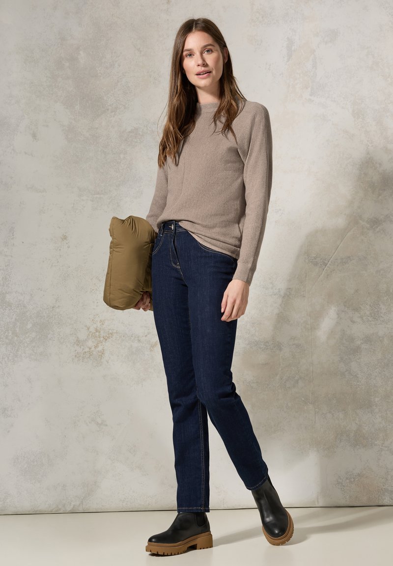 Maglione a maglia beige con maniche lunghe, jeans blu scuro e stivali neri alla caviglia con una suola spessa color cuoio. Tiene un giubbotto imbottito verde.