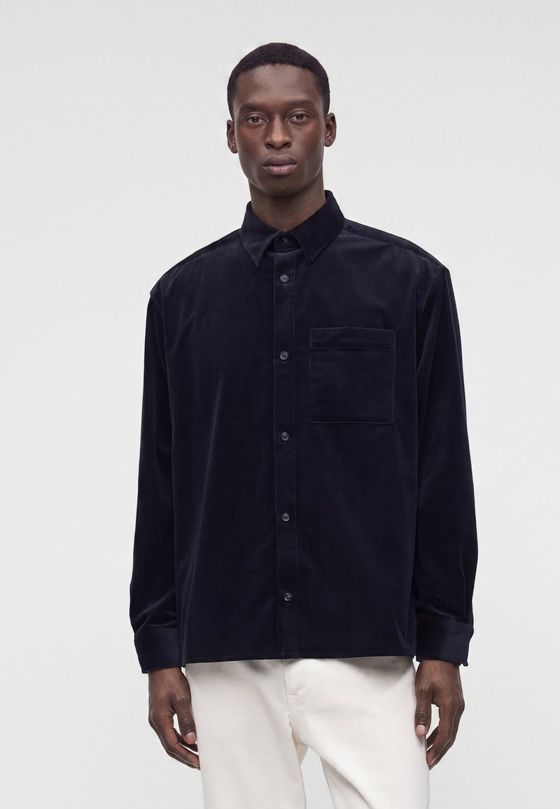 Homme portant une chemise noire à manches longues boutonnée avec une poche poitrine et un pantalon blanc, debout devant un fond clair uni.