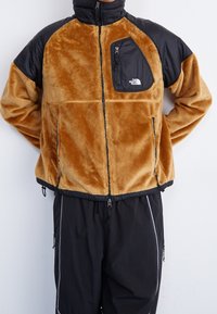 Giacca in pelliccia sintetica color sabbia con dettagli in nylon nero, tasca sul petto con zip, zip frontale, orlo elasticizzato e chiusure laterali. Abbinata a pantaloni neri.