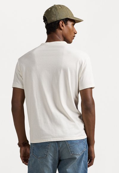 Balta kokvilnas t-krekls ar īsām piedurknēm, redzams no aizmugures, savienots ar ziliem džinsu biksēm un olīvu zaļu cepuri.