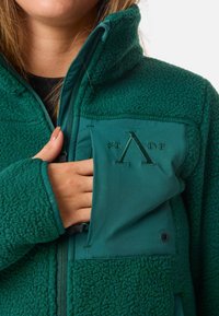 Giacca in fleece verde con finitura morbida e testurizzata e dettagli in tessuto liscio. Include una zip davanti e un logo ben visibile sulla tasca.