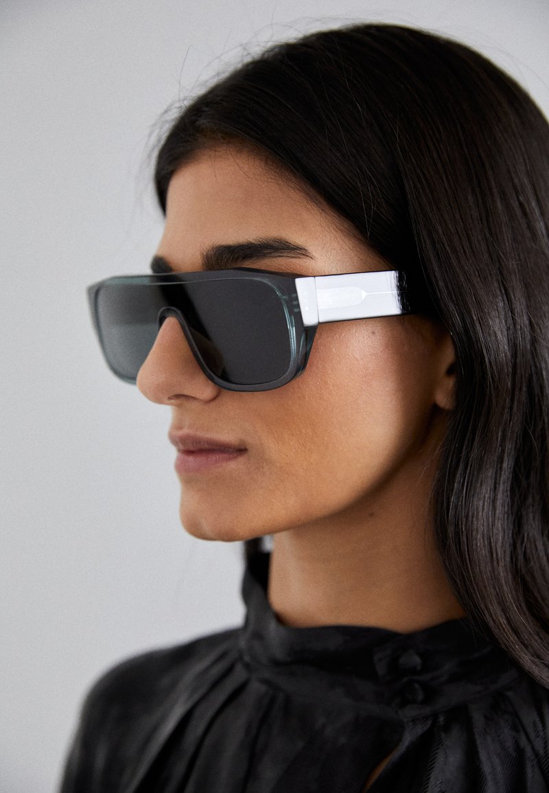 Occhiali da sole oversize con lenti scure, montatura trasparente grigia e accenti bianchi sugli steli; design angolare con una finitura lucida.