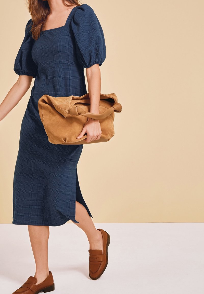 Femme en robe bleu marine à manches bouffantes tient un grand sac à main en suède marron clair, porte des mocassins marron, debout sur un fond beige et blanc.