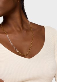 Naszyjnik z metalowego łańcucha w kolorze złotym i srebrnym z naprzemiennie ułożonymi owalnymi ogniwami, noszony na jasnobeżowej bluzce z prążkowanego materiału z głębokim dekoltem w kształcie V.