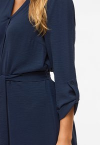 Γυναίκα φοράει μια μακρυμάνικη navy μπλε φόρεμα με γυρισμένα μανίκια και ύφασμα ζώνη δεμένη στη μέση, μαλλιά που ακουμπούν στον ώμο.