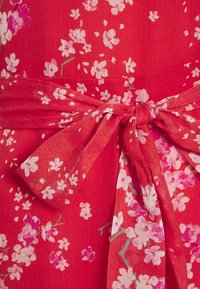 Červené květinové šaty s texturovanou tkaninou. Vyznačují se růžovými květinovými vzory, detaily uvázaného pásku a jemnou, splývavou kvalitou.