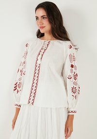 Blouse blanche avec manches longues ornées de broderies florales rouges, un accent central rouge et un col froncé pour plus de texture.