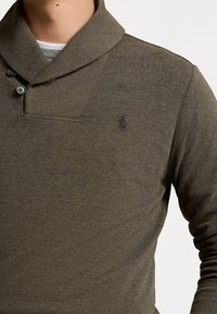 Homme portant un polo à manches longues vert olive avec col châle et boutons, superposé à un haut à col rond gris et blanc.