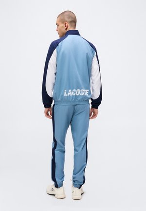 Mann trägt hellblauen, marineblauen und weißen Trainingsanzug mit dem Schriftzug „LACOSTE“ auf dem Rücken, steht mit dem Rücken zum Betrachter vor einfarbigem Hintergrund.