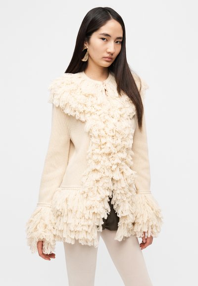 Κρεμ πλεκτή ζακέτα με υφασμάτινη, με φουντίτσες λεπτομέρεια στους ώμους και στο τελείωμα. Διαθέτει ριμπ μανίκια και στρογγυλή λαιμόκοψη.