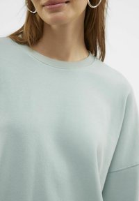 Lys grøn sweatshirt med ribbet rund hals og dropped shoulders, lavet af blødt bomuldsstof; har en glat tekstur og minimal design.