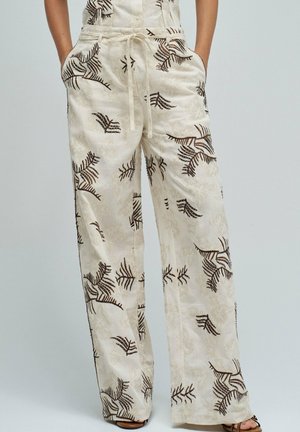 Pantalones de tela cremosa con cordón y piernas anchas, con patrones de hojas marrón oscuro, llevados con las manos en los bolsillos sobre un fondo liso.