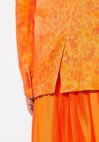 Blusa naranja con un patrón de estampado de leopardo, con una falda naranja plisada debajo. La blusa tiene botones redondos en los puños.