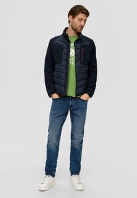 Giacca imbottita blu navy con dettagli in pile, tasche con zip e colletto alto, indossata sopra una t-shirt grafica verde, abbinata a jeans blu e sneakers bianche.