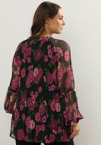 Musta läpikuultava blouse, jossa on vaaleanpunaisia ja vihreitä kukkakuvioita, päällä laskostettu rakenne, pitkät puffihihat, jossa on röyhelökauluksen yksityiskohtia, virtaava muotoilu.