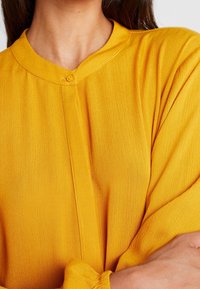 Gelbe Bluse mit strukturierter Oberfläche, runder Kragen, Knopfverschluss und gerafften Ärmeln für zusätzliche Details.