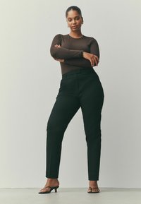 Haut marron à manches longues associé à un pantalon noir sur mesure et des sandales à talons noires. Le mannequin se tient les bras croisés, mettant en valeur la tenue.