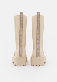 Botas de imitación de cuero beige hasta la rodilla con suela de plataforma gruesa y tirador con logotipo. Textura suave con detalles de costura visibles.