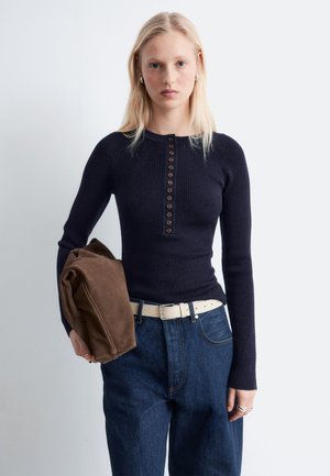 Vrouw met lang blond haar die een zwart vest met knopen draagt, blauwe jeans met een witte riem, en een opgevouwen bruine jas vasthoudt.