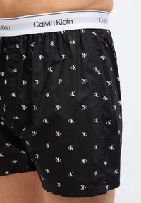 Μαύρα μποξεράκια Calvin Klein με λευκά λογότυπα "cK" και λευκή μέση με το κείμενο "Calvin Klein", φορεμένα στο κάτω μέρος του κορμού ενός ατόμου.