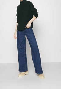 Donkergroene gebreide coltrui, lichtblauwe jeans met wijde pijpen en beige chunky-solelaarzen. Gladde textuur, ontspannen pasvorm en rechte silhouet.