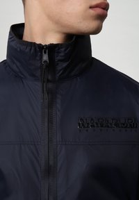 La giacca zip-up blu navy presenta un colletto alto, un tessuto testurizzato e un logo in gomma nera sul lato sinistro del petto, con il cursore della cerniera visibile.