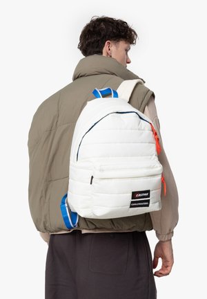 Mochila acolchada blanca con correas azules y tiradores de cremallera naranjas. Cuenta con un bolsillo delantero y una etiqueta de marca. Lleva puesta sobre una chaqueta acolchada gris.